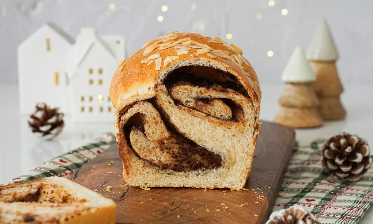 Коледно руло с меко тесто със златиста коричка и вихри от тъмен шоколад, поръсено с филирани бадеми върху дървена дъска
