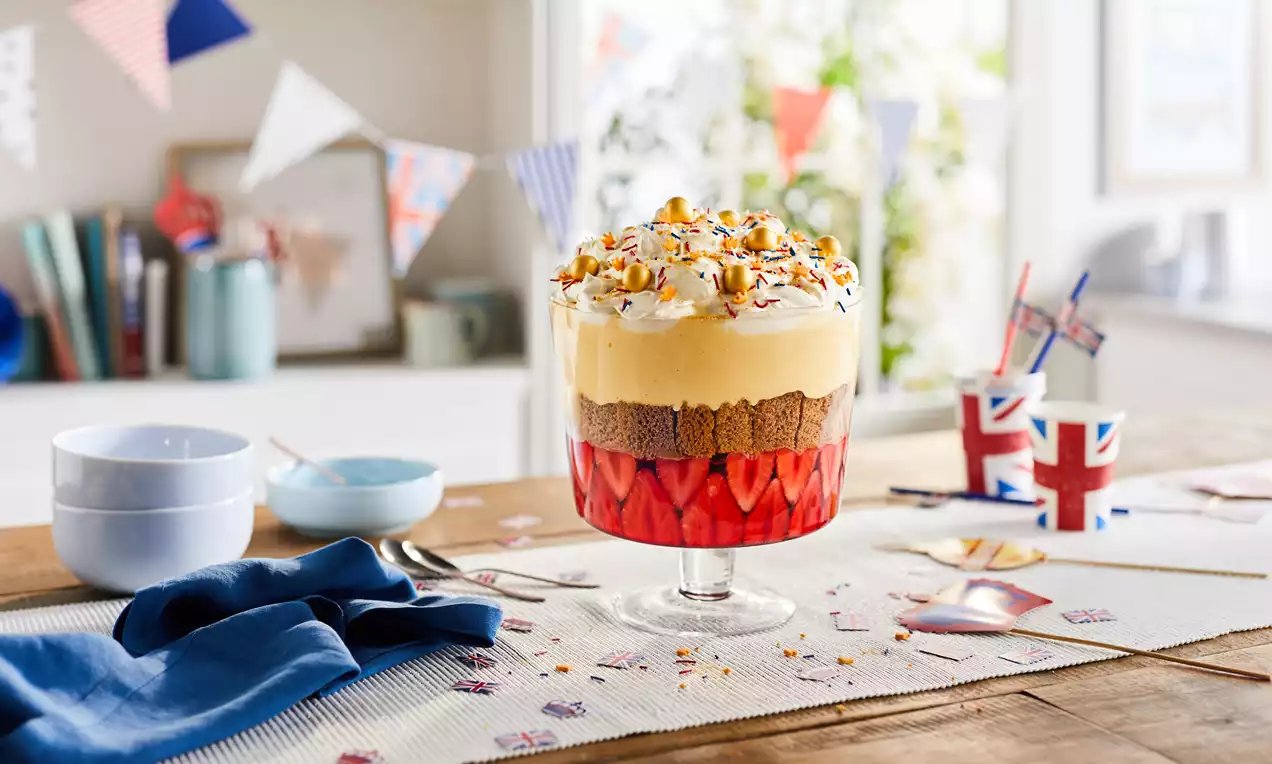 Layered Coronation trifle in a clear glass bowl with vibrant red strawberries, golden sponge, creamy custard, whipped cream, and sprinkles