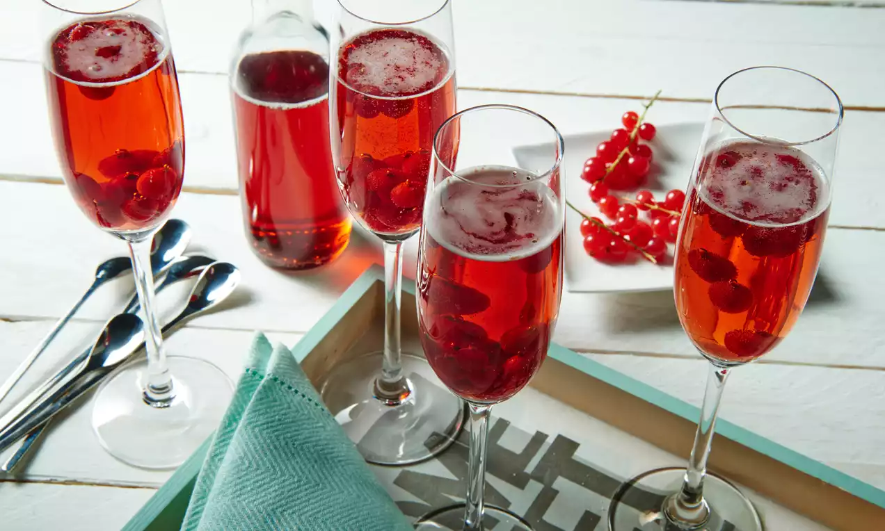 Gläser mit rotem Johannisbeer-Sirup, schwimmenden Beeren und Schaumkrone, arrangiert mit Flasche, Löffeln, Serviette und Johannisbeerrispen