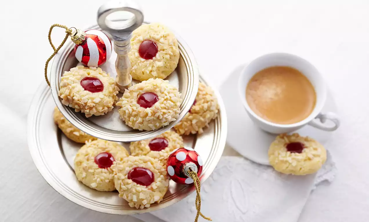 Kulleraugen mit goldgelbem Teig, gehackten Mandeln und roter Marmeladenfüllung, auf einer silbernen Etagere neben einer Tasse Kaffee drapiert