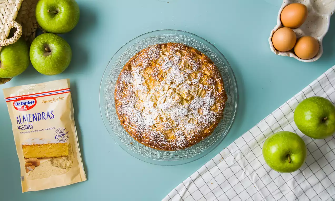 Tarta redonda de manzana con corteza dorada y superficie espolvoreada con azúcar glas sobre un plato de cristal, rodeada de manzanas verdes y huevos