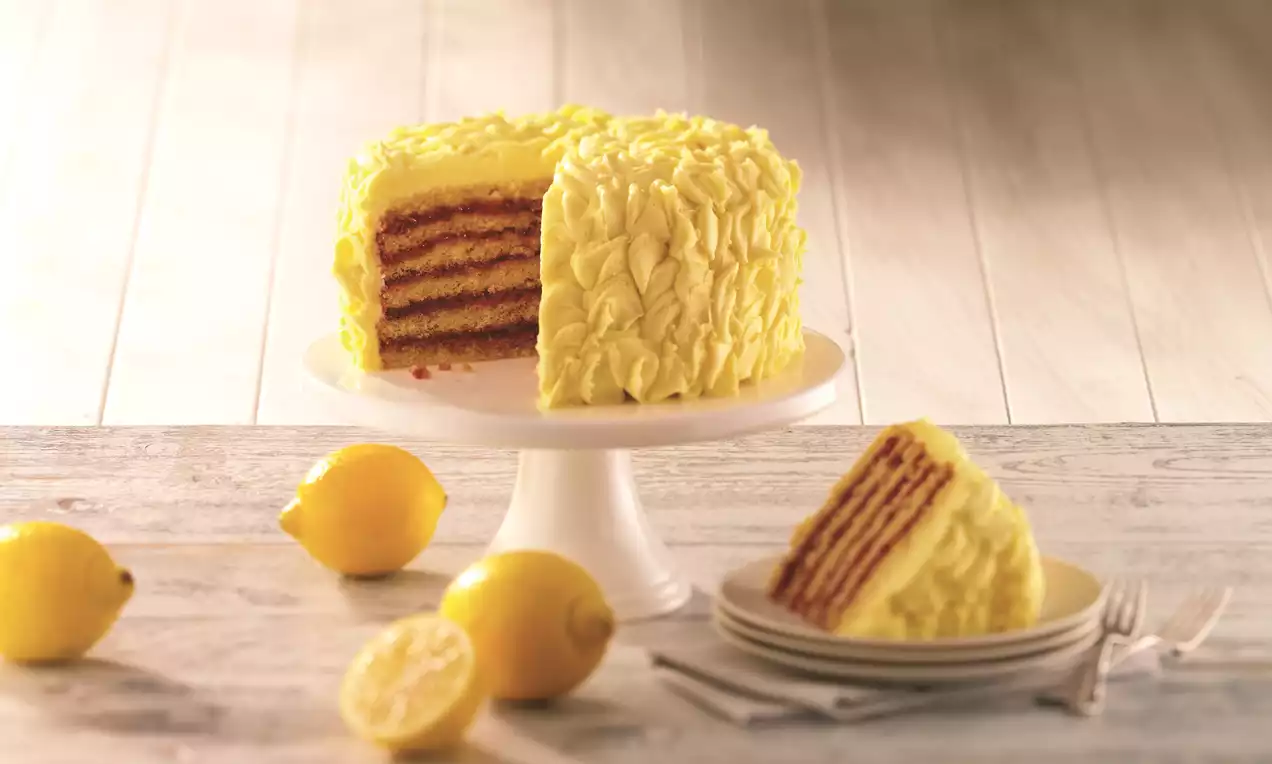 A tall layered cake with rich raspberry filling and vivid yellow ruffled frosting on a white pedestal, surrounded by fresh lemons