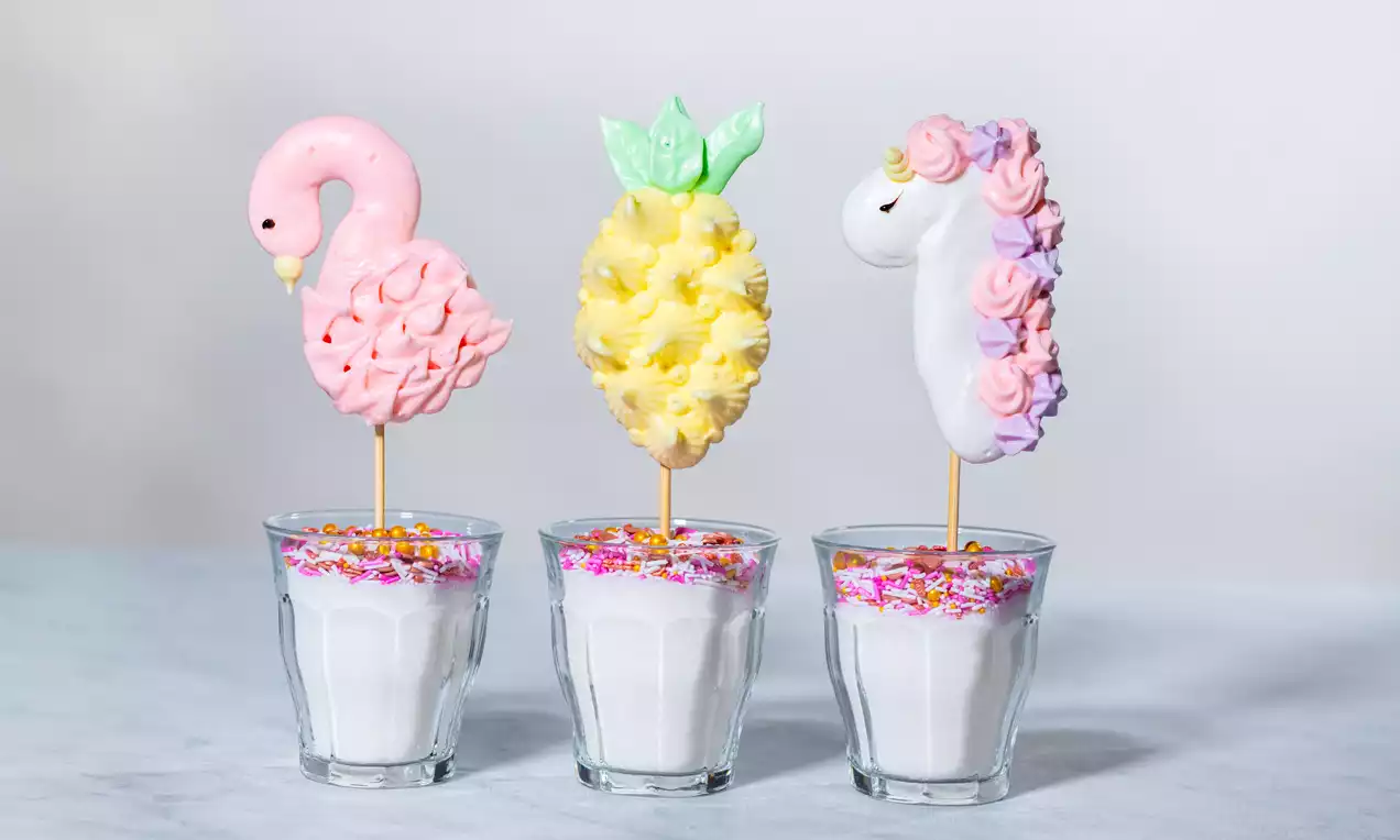 Bunte Meringue-Lollipops in Flamingo-, Ananas- und Einhorn-Form stecken in Gläsern mit weißem Zuckerguss und bunten Streuseln
