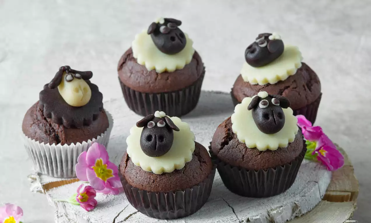 Kakaové muffiny s čokoládovými ovečkami na krémové polevě, uspořádané na rustikálním dřevěném podnosu s růžovými kvítky okolo