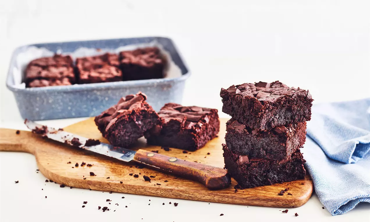 Carrés de brownie chocolatés denses et moelleux aux coins nets empilés sur une planche en bois avec un couteau et un linge bleu