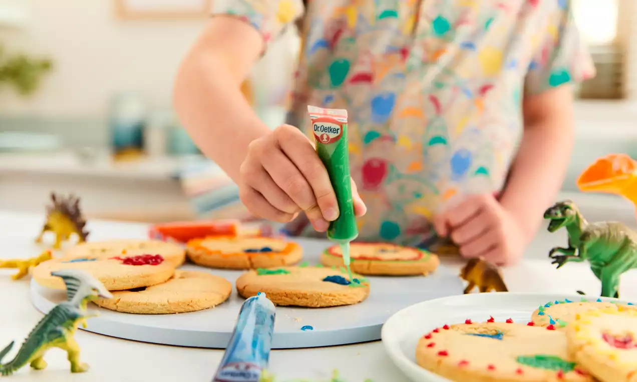 Golden round cookies with smooth surfaces being decorated with green icing, surrounded by toy dinosaurs on a white plate