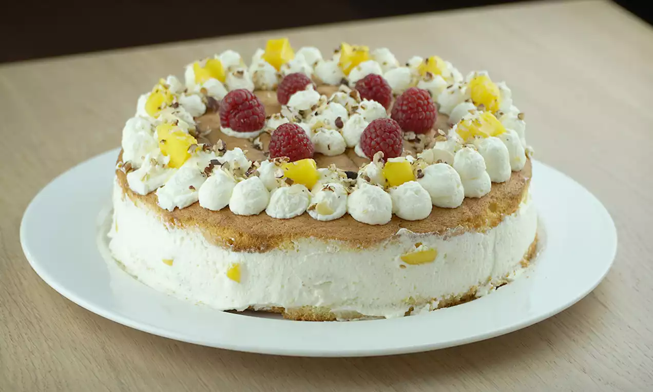 Gâteau à la crème blanche avec des morceaux de mangue, garni de framboises rouges, noisettes concassées et choux de crème sur une assiette blanche