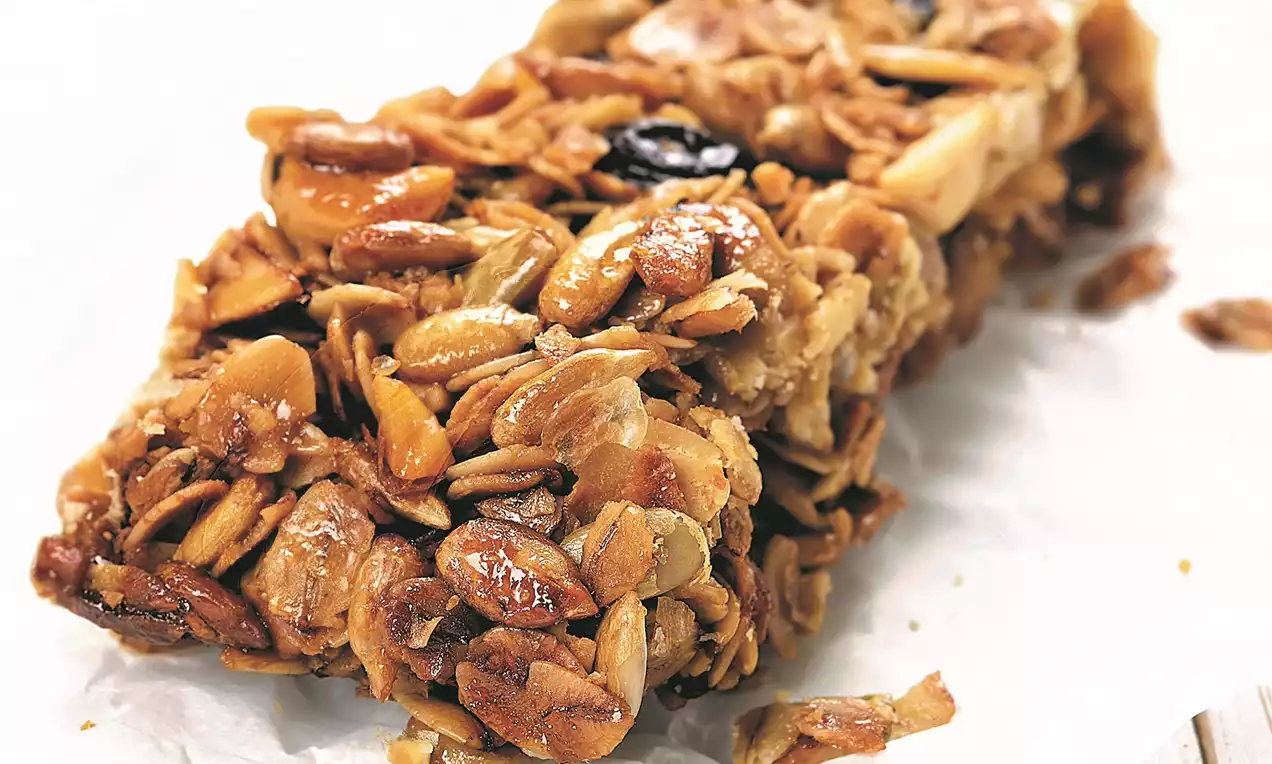 Golden brown granola bar packed with clusters of oats, nuts, and seeds, with a glossy finish resting on crinkled white parchment paper