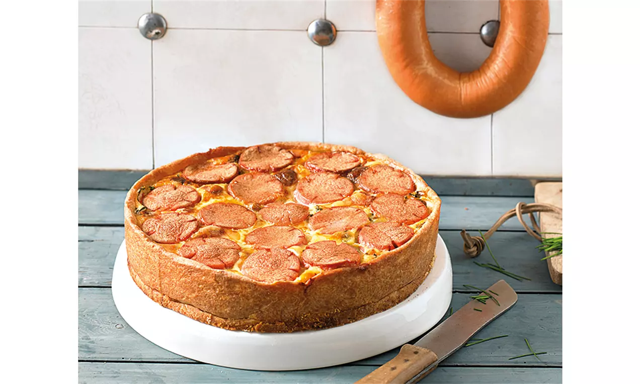 Goldbraune Lyonertorte mit knusprigem Teigrand, dichten Wurstscheiben und cremiger Füllung auf weißer Platte, angerichtet auf Holzbrett