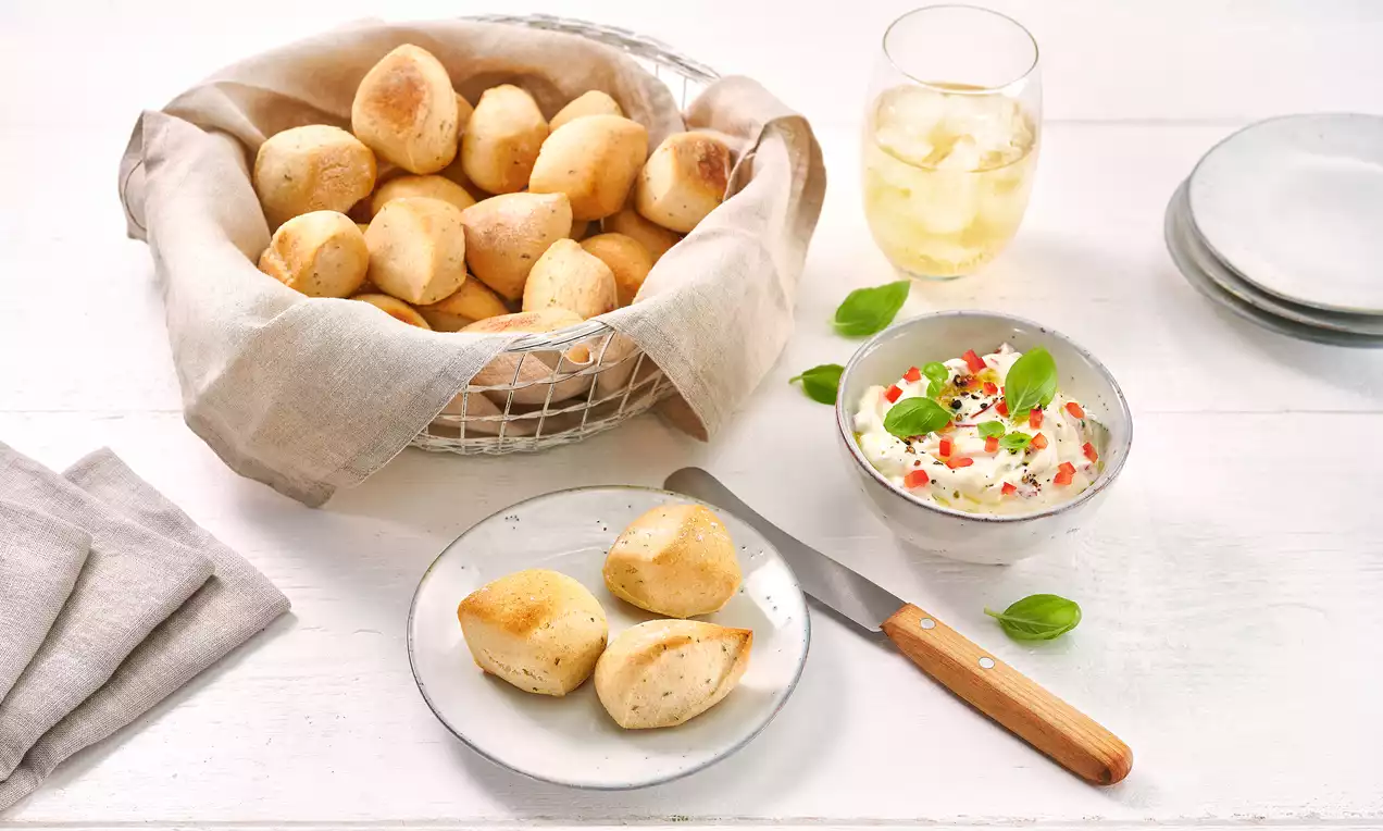 Goldbraune, runde Pizzabällchen in einem Stoff ausgelegten Korb und auf einem Teller, daneben Kräuter-Dip mit Basilikum und Getränk