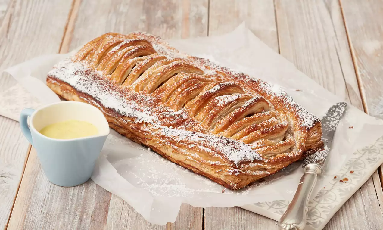 Goldbrauner Apfelstrudel aus Blätterteig mit sichtbaren Teigschichten, Puderzucker bestäubt auf Backpapier, daneben Vanillesoße in blauer Kanne