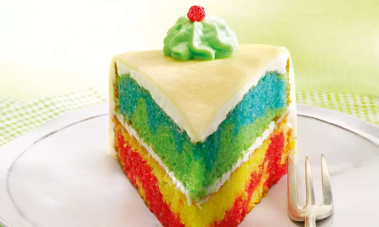 Dreieckiges Kuchenstück mit gelbem Überzug, bunter Schichtung in rot, gelb, grün und blau, dekoriert mit grünem Sahnetupfen und roter Perle