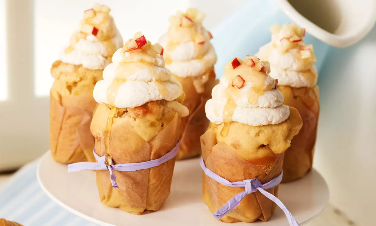 Cupcakesi zlatnožute nijanse, omotani papirom s ljubičastim trakama, s vrhom od bijelog šlaga, posuti kockicama crvenih jabuka i preliveni medom