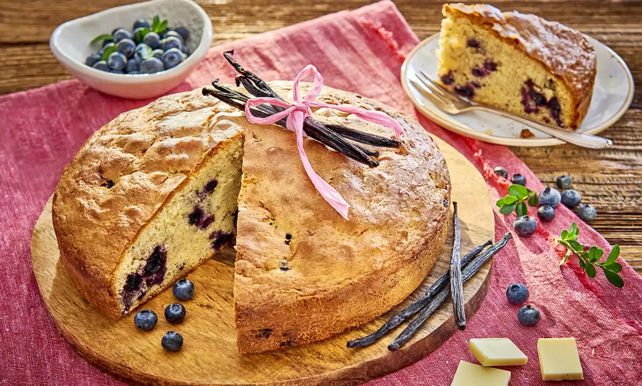 Okrągłe ciasto o złocistej skórce, z jagodami zatopionymi w jasnym wnętrzu, udekorowane laskami wanilii i różową wstążką na drewnianej desce