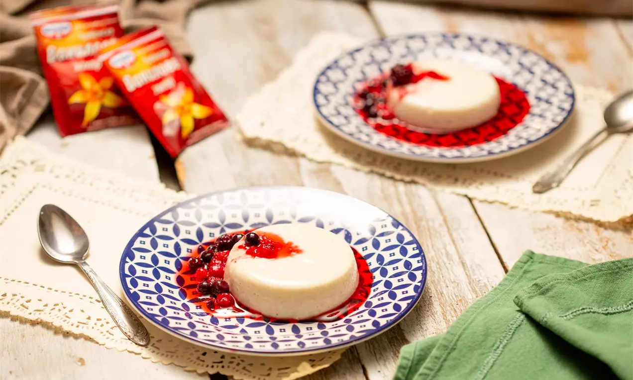 Панакотата с мек кремав оттенък е гладка и кръгла, обвита в яркочервен сос с цели горски плодове върху бяла чиния с тъмносин орнамент