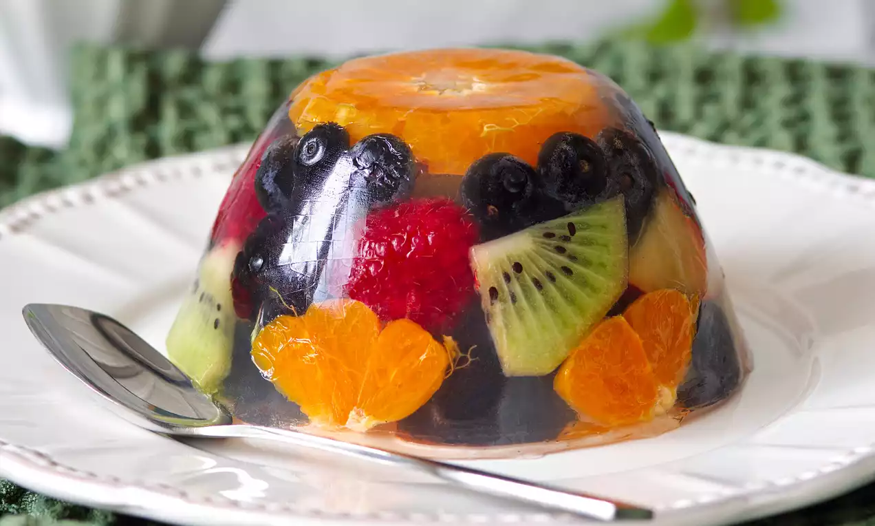 Barevné ovocné želé v průhledné kopulovité formě s plátky kiwi, mandarinek, malin a borůvek na bílém talíři s dezertní lžičkou