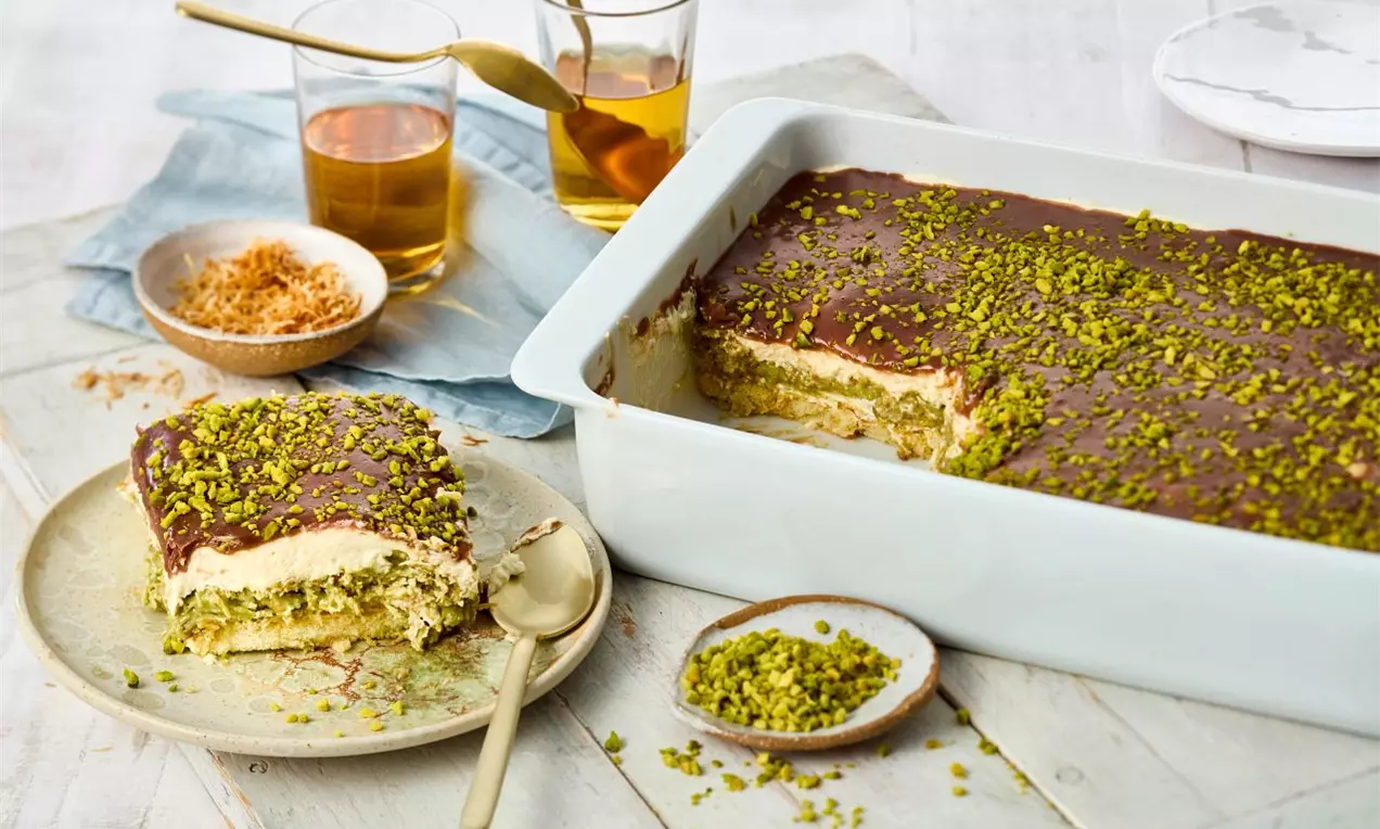 Čokoládové tiramisu s hladkou lesklou polevou, vrstvy světlého krému a pistácie na povrchu v bílé zapékací míse a na talíři s dezertní lžící