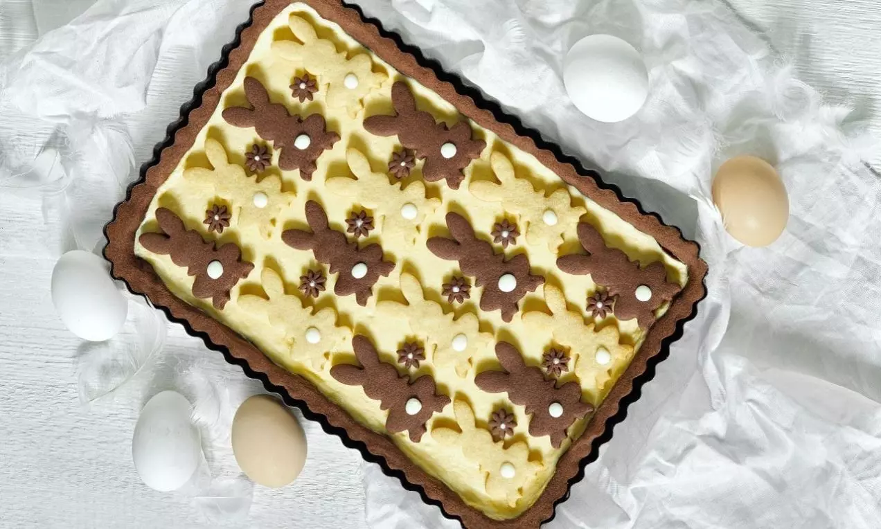 Tvarohový koláč na hnědém křupavém podkladu, se žlutou hladkou náplní, zdobený kakaovými zajíčky a květinami ve vzoru na bílém podkladu s vejci