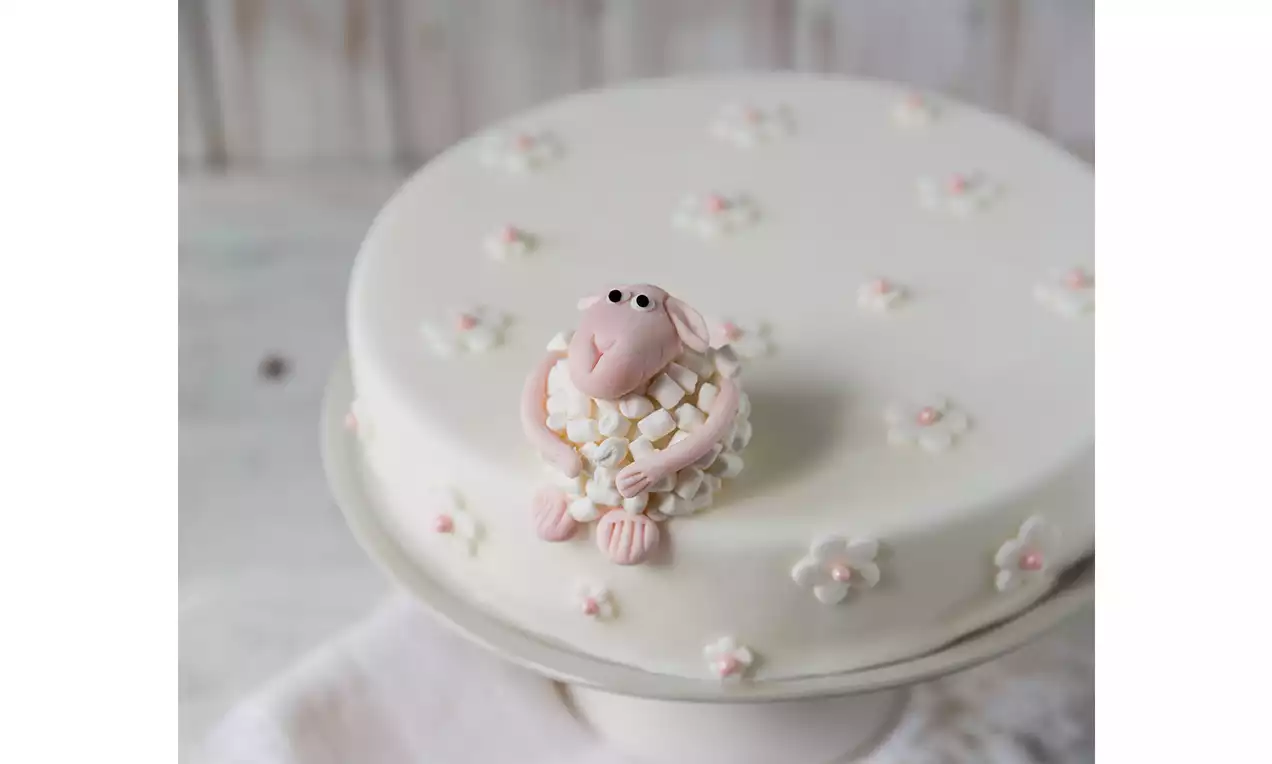 Agnellino in pasta di zucchero rosa con riccioli bianchi su torta al cioccolato ricoperta di glassa bianca decorata con piccoli fiori
