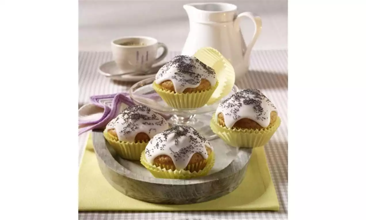 Golden muffins with smooth white cream glaze, sprinkled with dark poppy seeds, served in pale yellow cases on a wooden tray