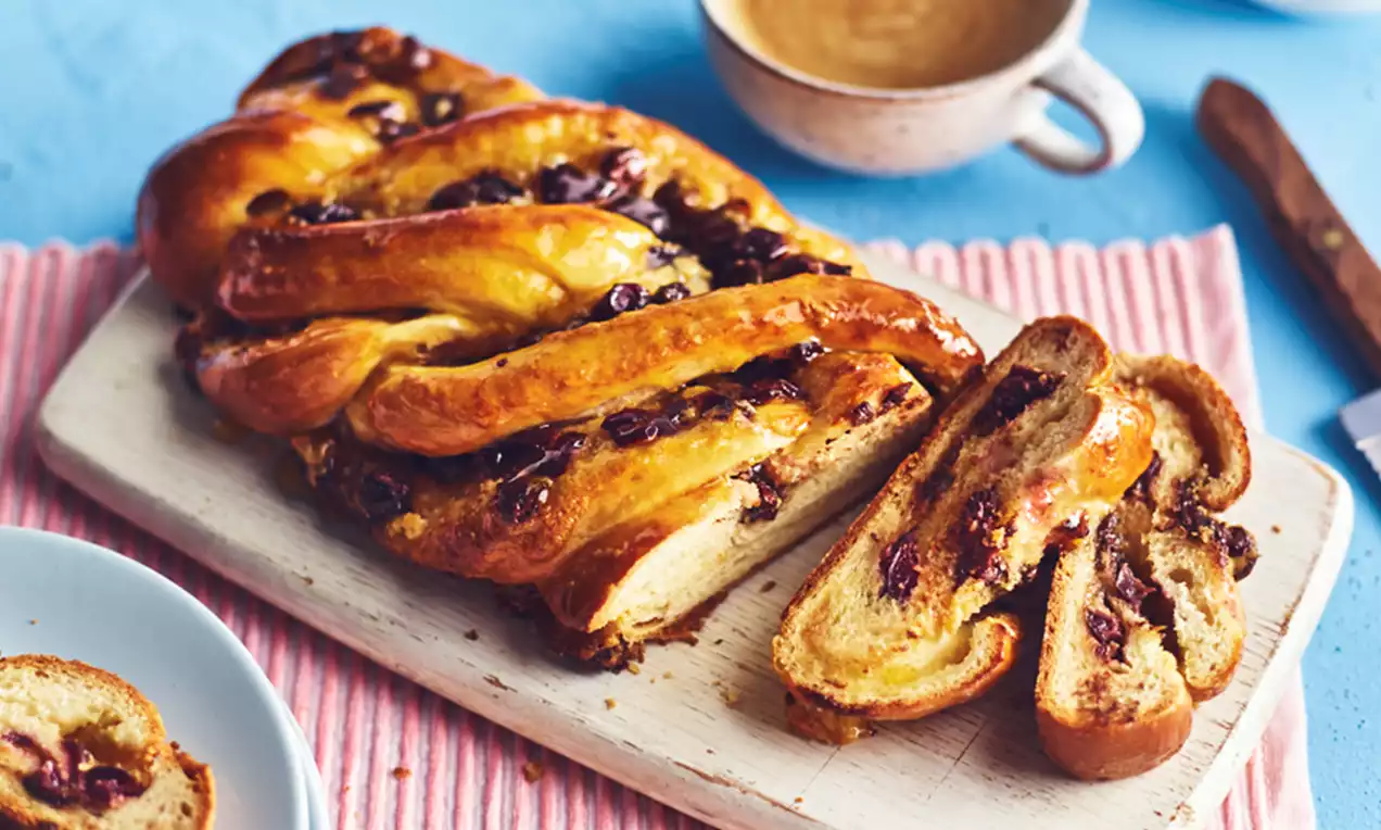 Golden braided pastry glistening with glaze, filled with dark cherries and chocolate, served sliced on a wooden board with a coffee cup nearby
