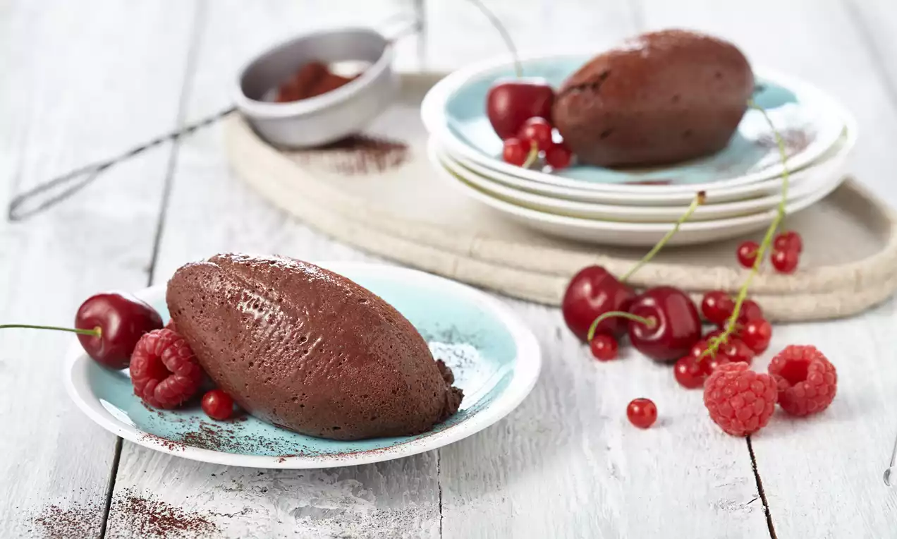 Schokoladen-Mousse als oval geformte Nocken mit glatter, dunkler Oberfläche, dekoriert mit Himbeeren, Kirschen und Kakao auf hellblauen Tellern