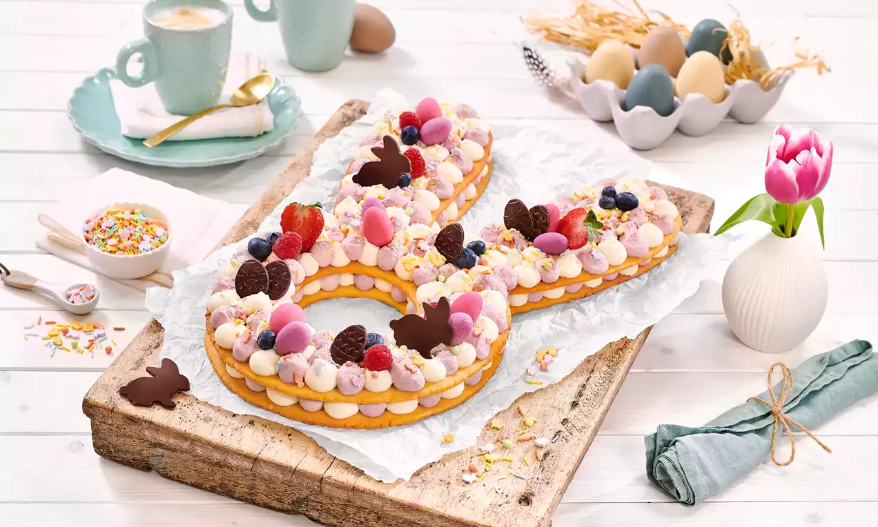 Letter cake en forme de lapin avec couches dorées, crème blanche, framboises rouges, œufs roses et violets et papillons chocolat sur planche