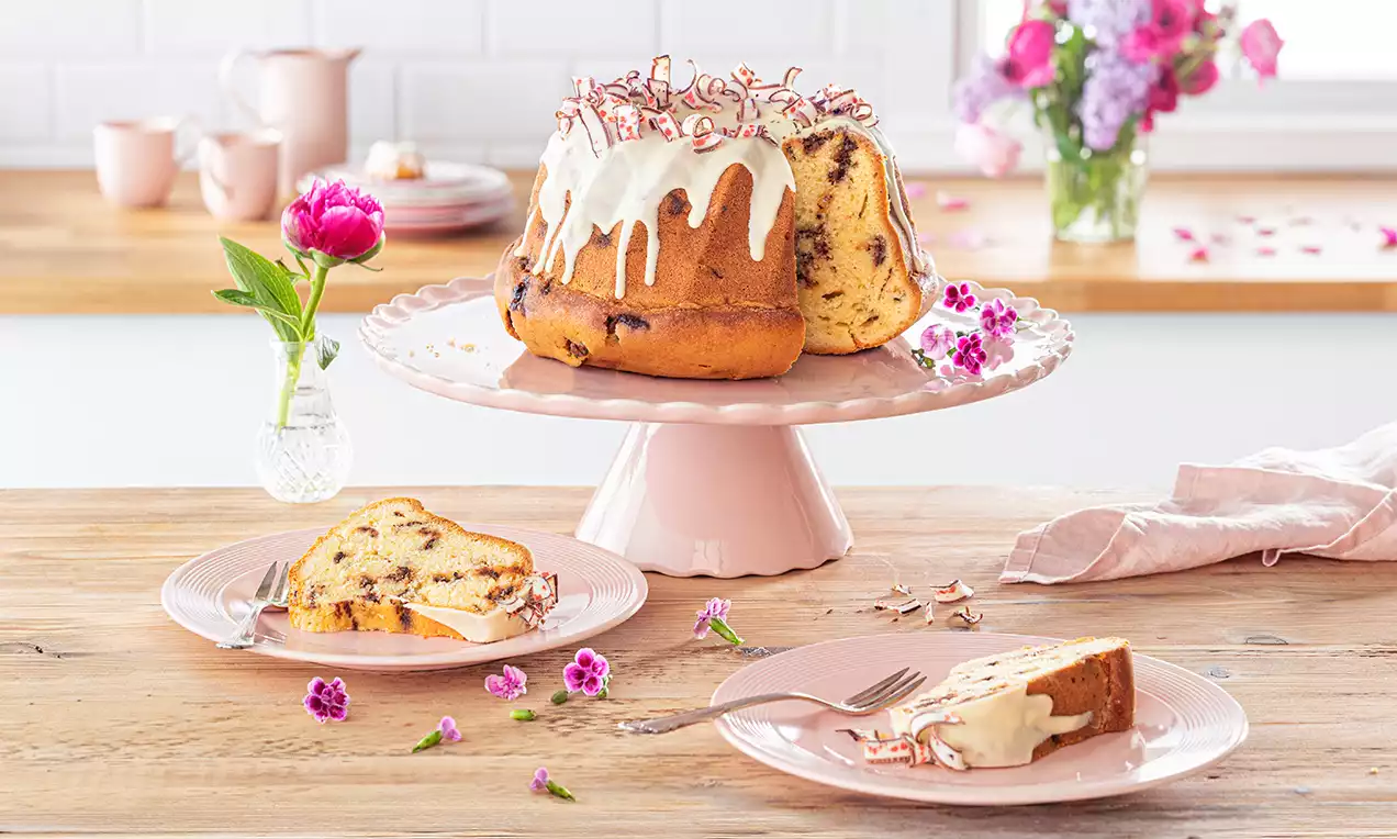 Erdbeer-Joghurt-Gugelhupf mit heller Glasur und Schokostückchen auf rosa Tortenplatte, rosa Teller mit angeschnittenem Stück und Blütendeko