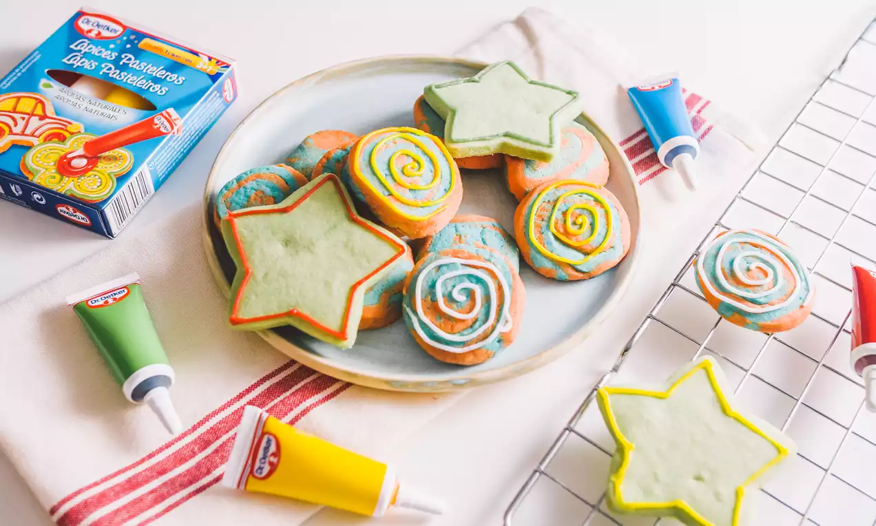 Galletas en espiral de colores vivos y formas de estrella, decoradas con glaseado brillante, dispuestas sobre un plato blanco y mantel rayado