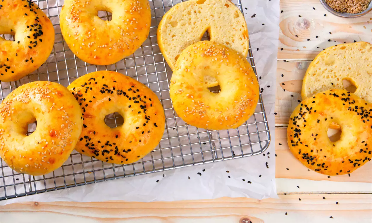 Goldbraune Protein-Bagels auf einem Abkühlgitter, bestreut mit Sesam und Schwarzkümmel. Daneben halbierte Bagels auf einem Holztisch sowie verstreute Körner. Helles, freundliches Setting.