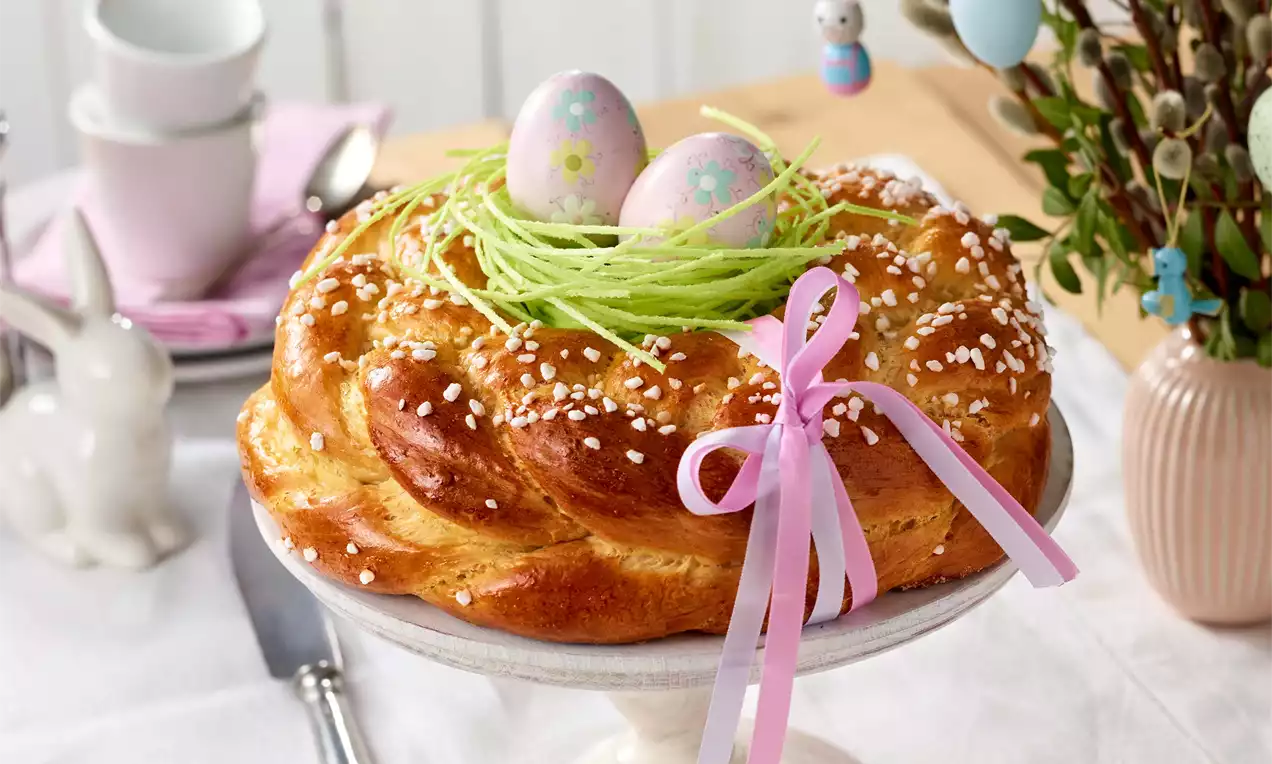 Zlatistý vrkoč posypaný hrubým kryštálovým cukrom, uprostred s hniezdom zo zelených prúžkov a dvoma ružovkastými veľkonočnými vajíčkami