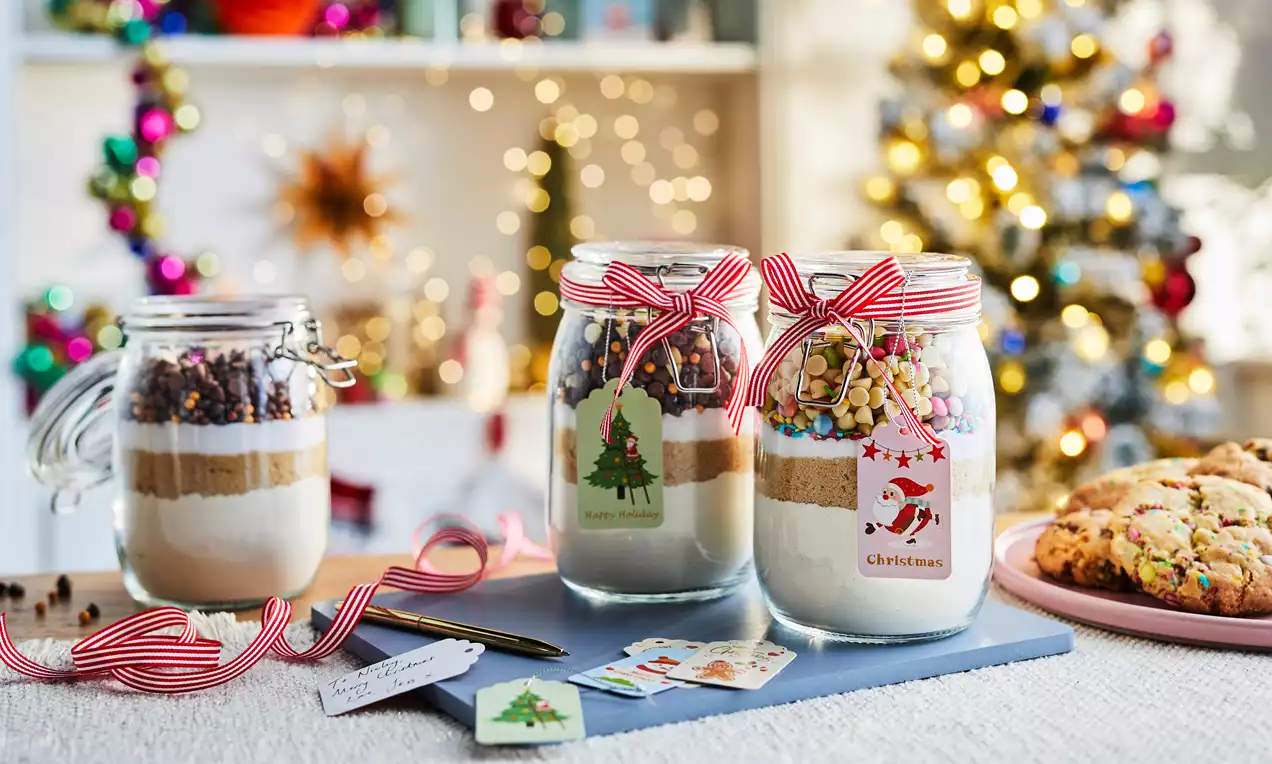 Glass jars layered with white flour, brown sugar, and chocolate chips, topped with festive ribbons and tags, set on a table with a blurred Christmas tree in the background