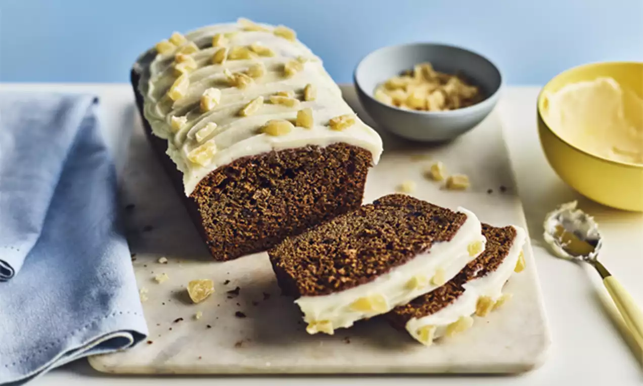 Dark brown ginger loaf cake with a moist texture, topped with white icing and chopped candied ginger, served on a marble board