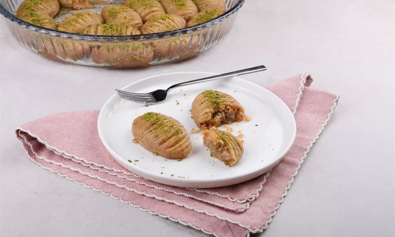 Oval biçimli ve altın rengi bıçak tatlıları, ince çizgili yüzeylere sahip, iç dolgusu açıkta, üzerine toz yeşil fıstık serpilmiş