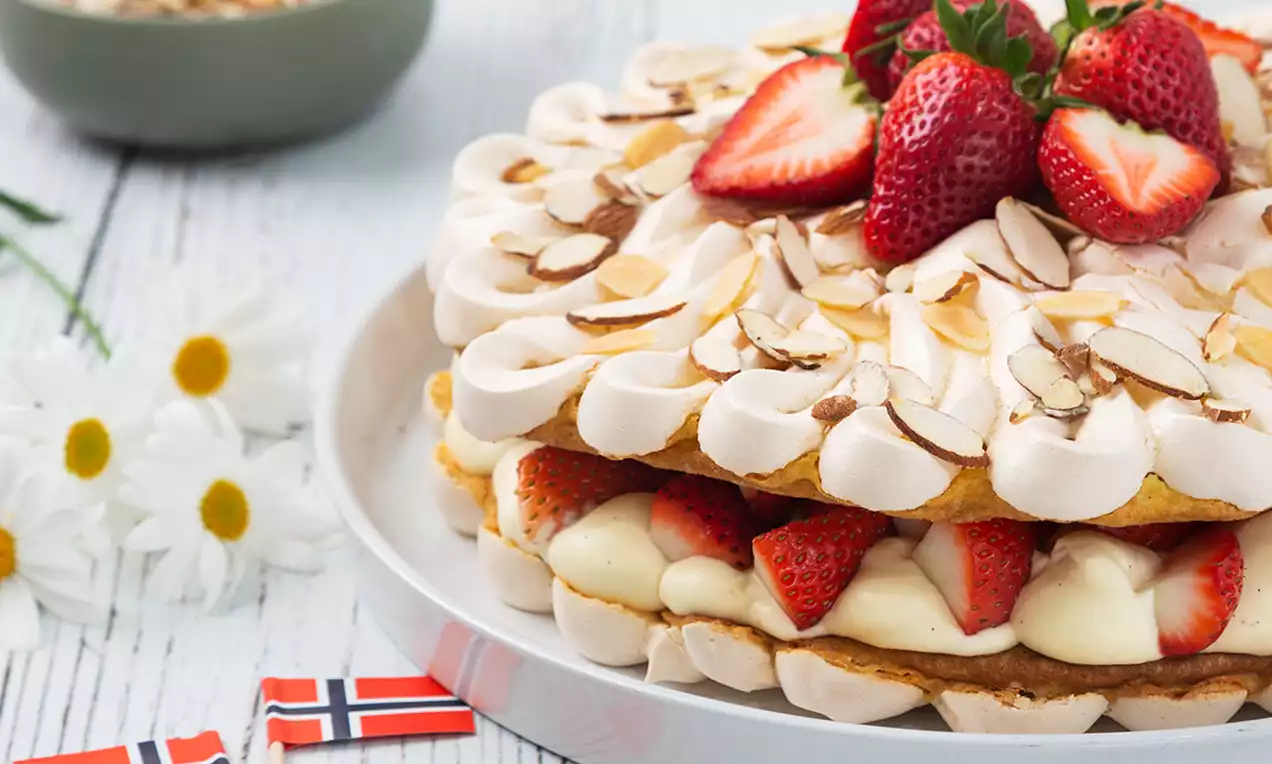 Kake i lag med krem, jordbær og kjeksbunn toppet med mandelflak og jordbær, servert på hvit tallerken med blomster og norske flagg