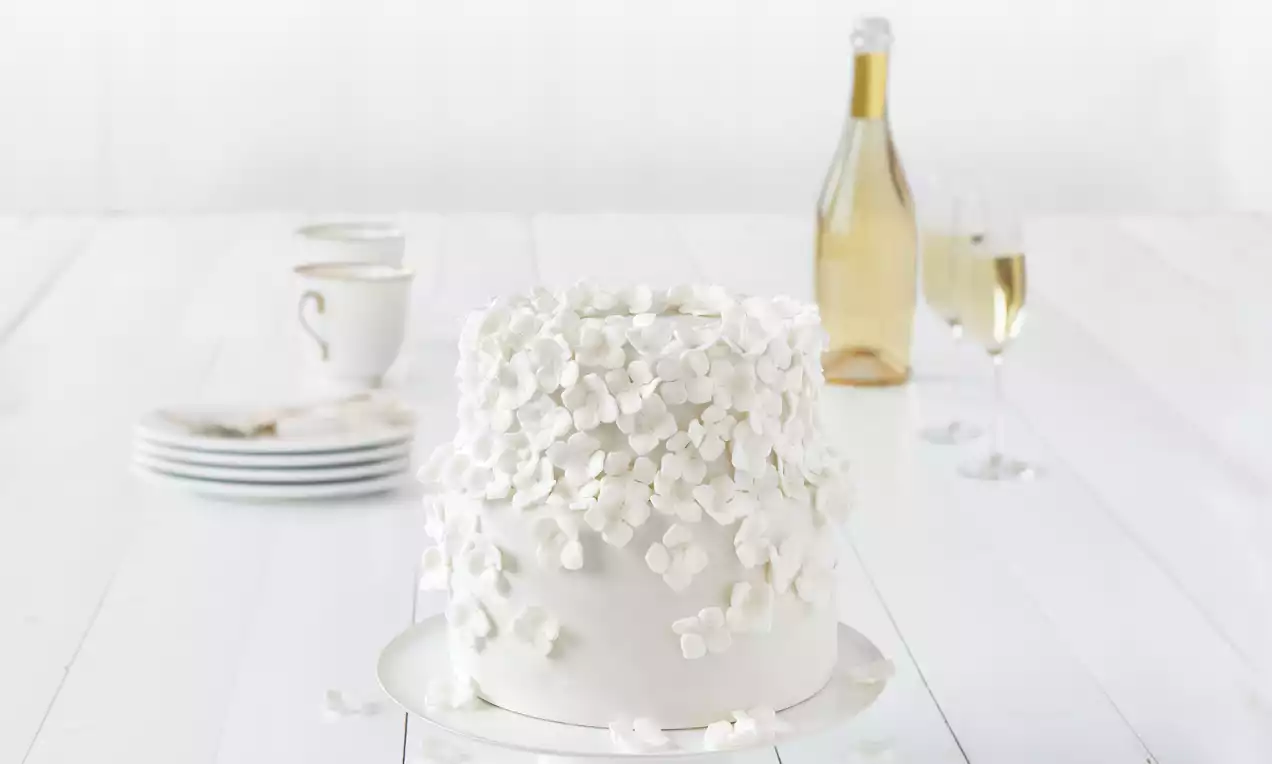 Lumivalkoinen kookos-limettikakku pyöreässä muodossa, sileän kuorrutteen pintaa koristavat ilmavat kukkalastut tasaisesti aseteltuna