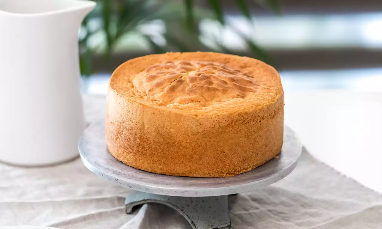 Bizcocho Genovés dorado y esponjoso con borde liso y superficie ligeramente abombada, sobre un plato metálico gris sobre mantel claro