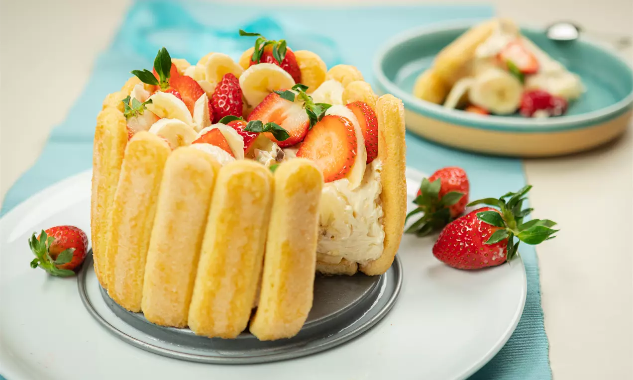 Банановата шарлота с меки бишкоти във форма на кръг, украсена с нарязани банани, ягоди и крем, лежи върху сива и бяла чиния