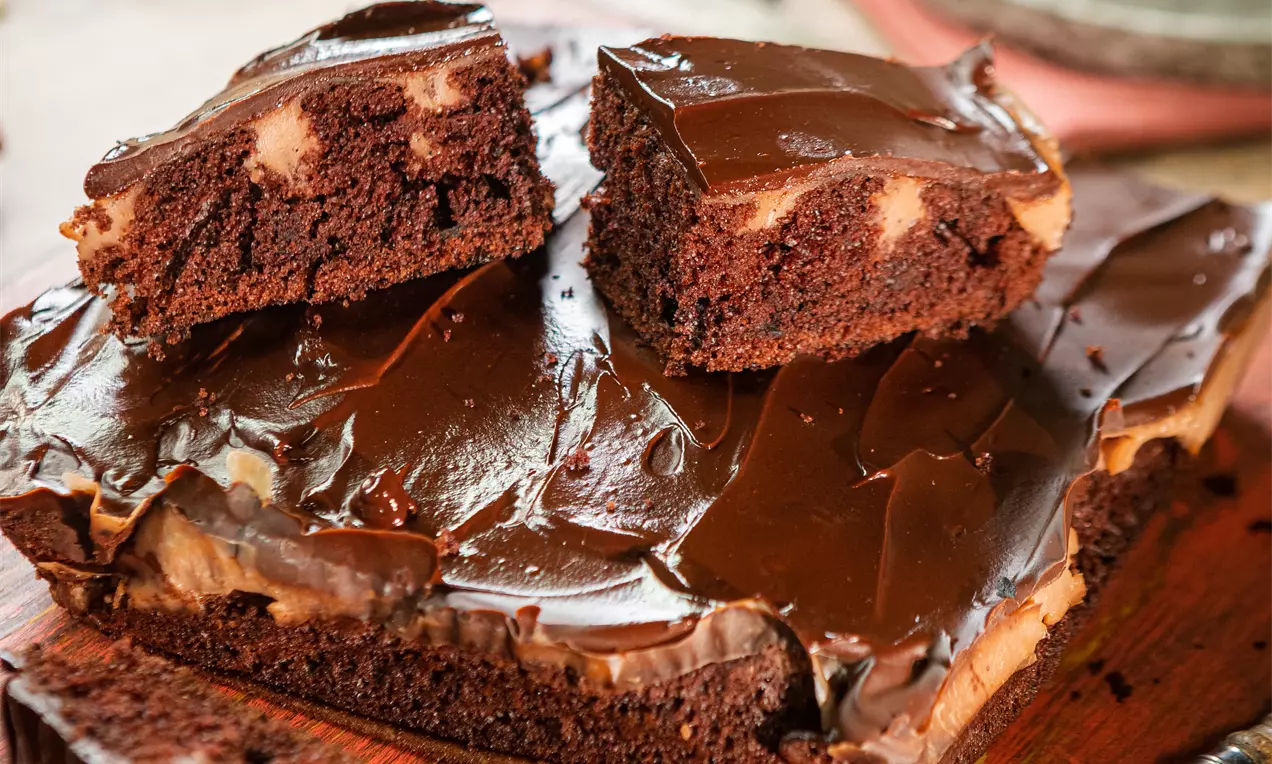 Кейк с плътен кафяв блат, покрит с гладка тъмночоколадова глазура, с мраморен ефект на парчета, поставен върху дървена дъска