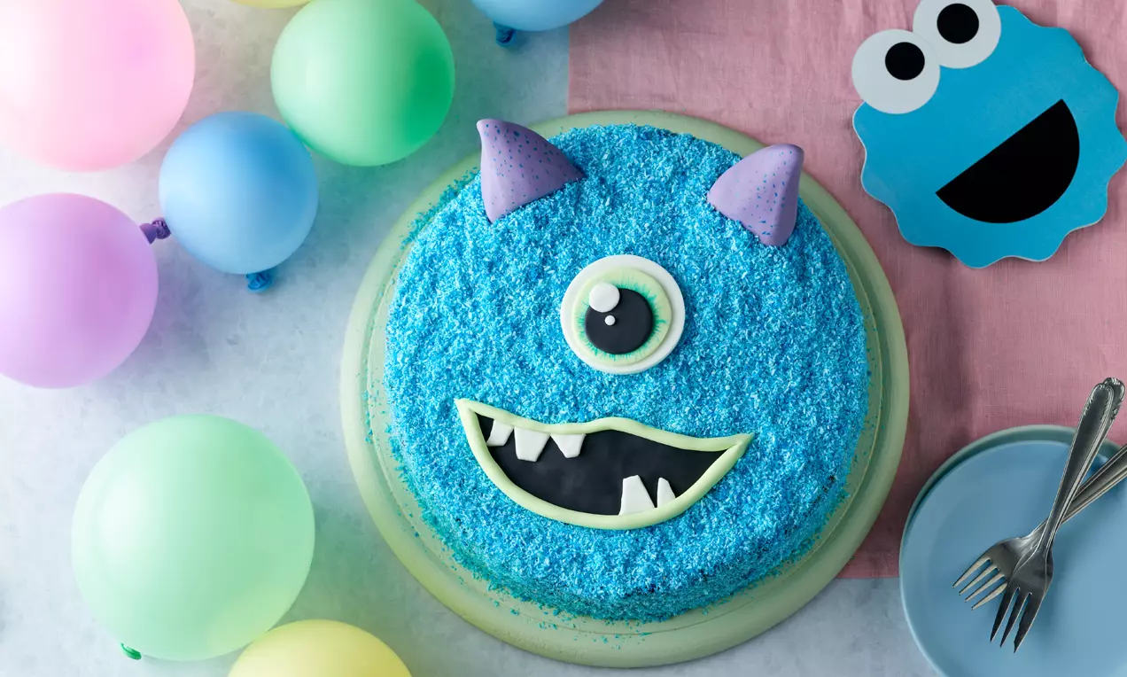 Runde Torte mit blauer Kokosraspel-Oberfläche, einem grünen Monsterauge, weißen Zähnen, lilafarbenen Hörnern, umgeben von bunten Ballons