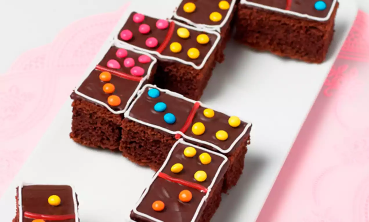 Brownies rectangulares de chocolate oscuro con glaseado brillante, líneas de azúcar blanca y puntos de colores dispuestos como fichas de dominó sobre un plato blanco
