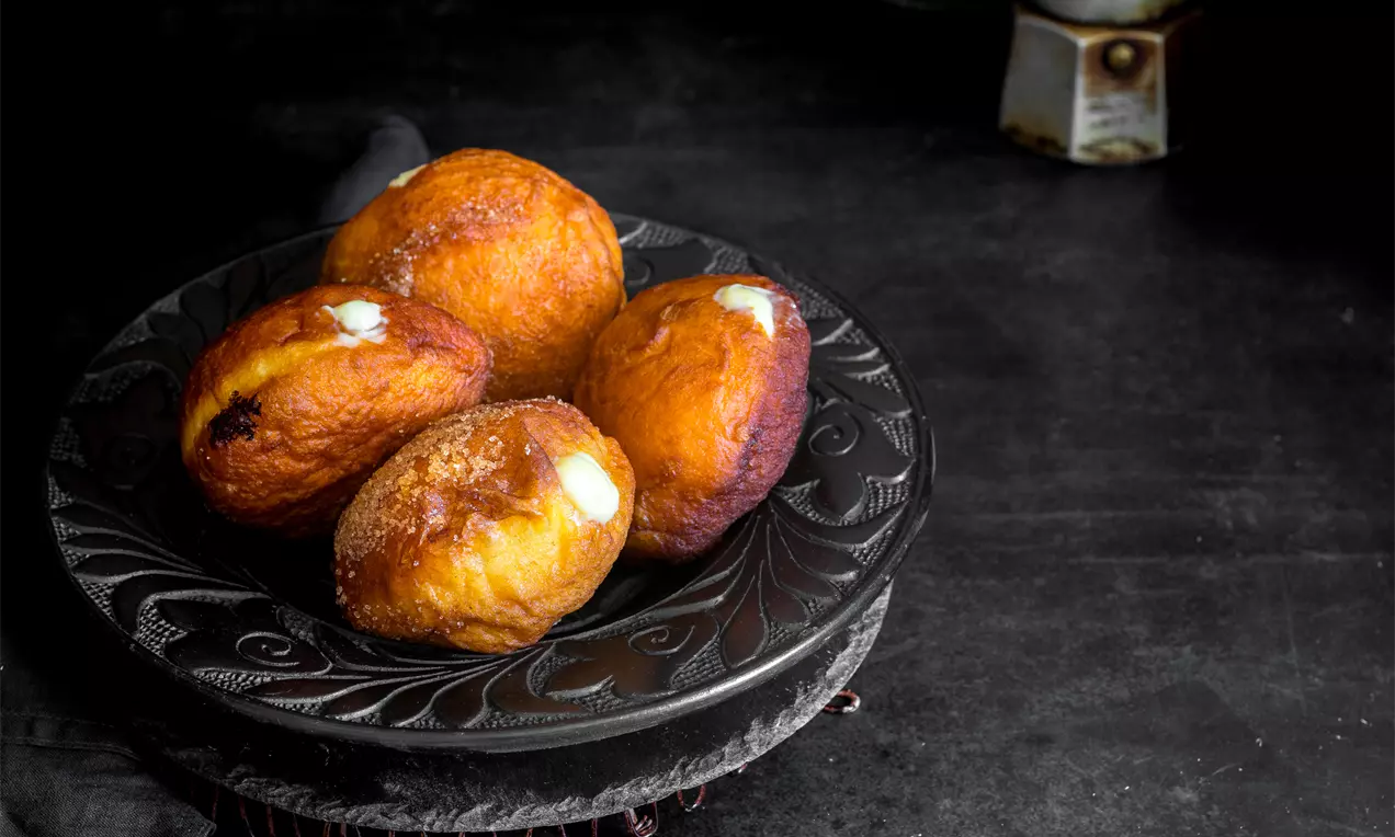 Златисто-кафяви донъти с пухкава текстура, плътно подредени върху тъмна декоративна чиния, с кремообразен пълнеж и поръсени със захар