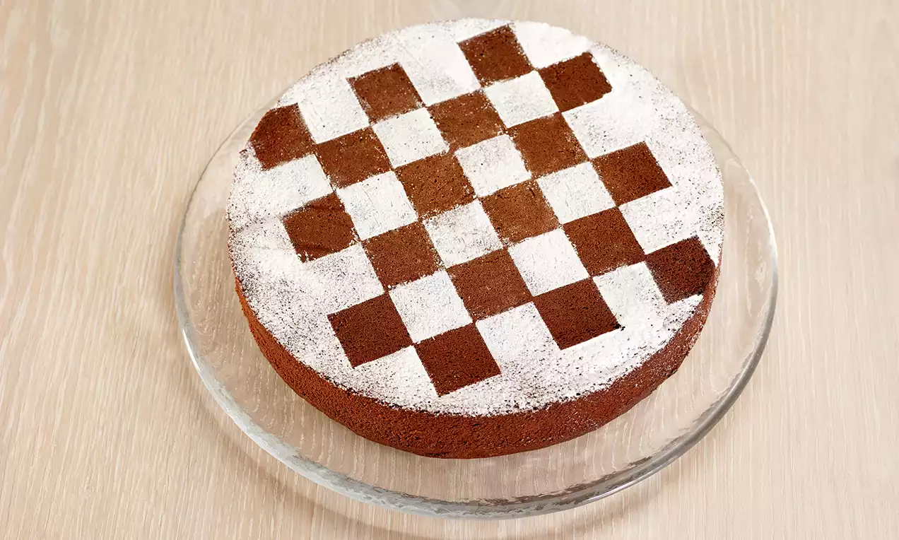Torta al cioccolato scura, soffice e rotonda con decorazione a scacchiera di cacao e zucchero a velo su un piatto trasparente di vetro