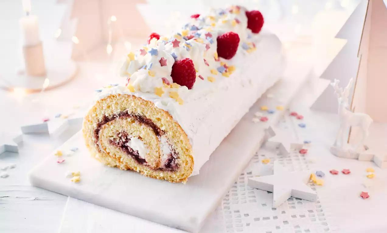 Bûche roulée à la génoise jaune pâle garnie de confiture rouge et crème blanche, décorée de framboises et étoiles colorées sur un plateau lumineux