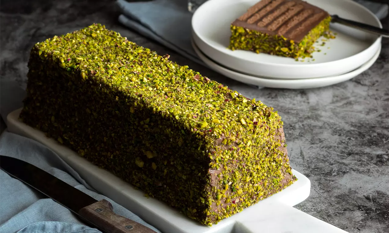 Правоъгълна шоколадова торта, покрита с плътен слой натрошени зелени шамфъстъци, сервирана върху бяла плоска чиния върху сив фон
