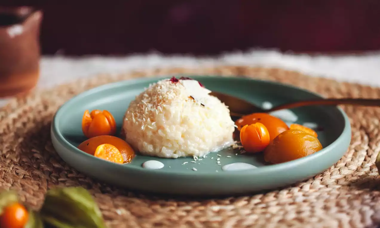 Koule lepkavé rýže s jemným kokosovým posypem, obklopená jasně oranžovými meruňkami a physalis na matně zeleném talíři