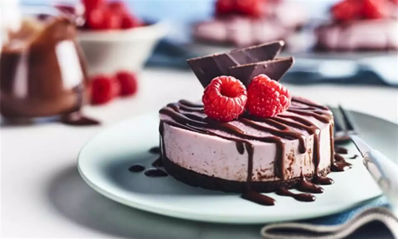 Cheesecake vegan cu strat roz-pal de cremă de zmeură, blat maro crocant, decorat cu glazură de ciocolată, zmeură proaspătă și bucăți de ciocolată pe o farfurie deschisă la culoare