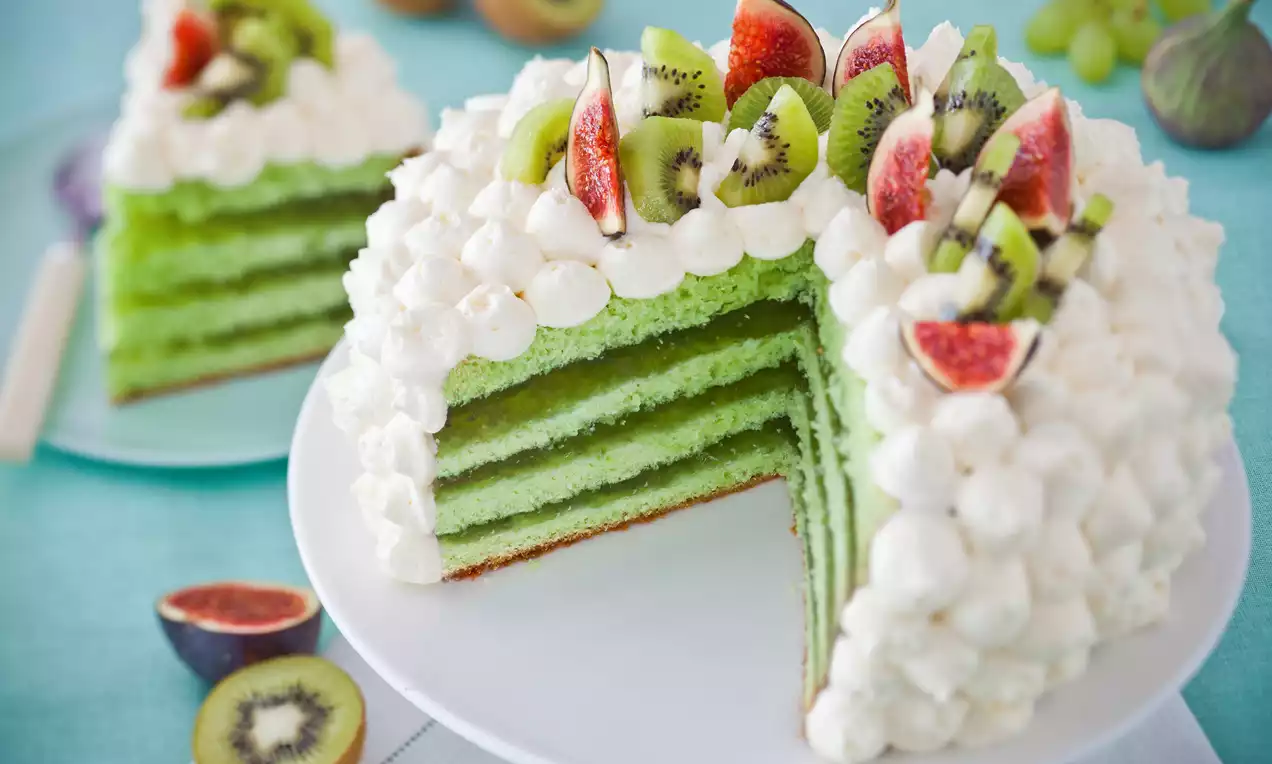 Tort warstwowy w odcieniach zieleni, ozdobiony białymi rozetkami kremu, plasterkami kiwi i fig na białym talerzu