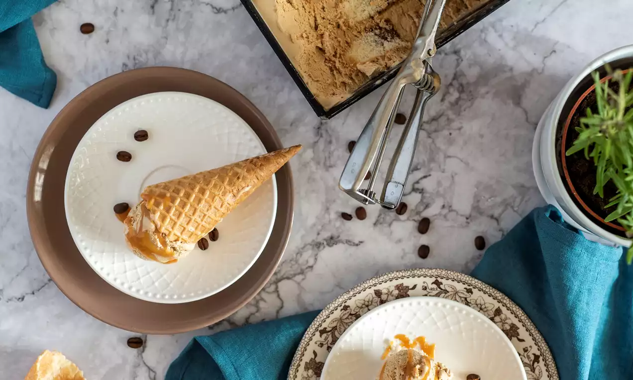 Tiramisu saldējums karameļu tonī, krēmīgs un viegli marmorēts, pasniegts kraukšķīgā vafeļu konusā uz porcelāna šķīvja ar kafijas pupiņu dekoru