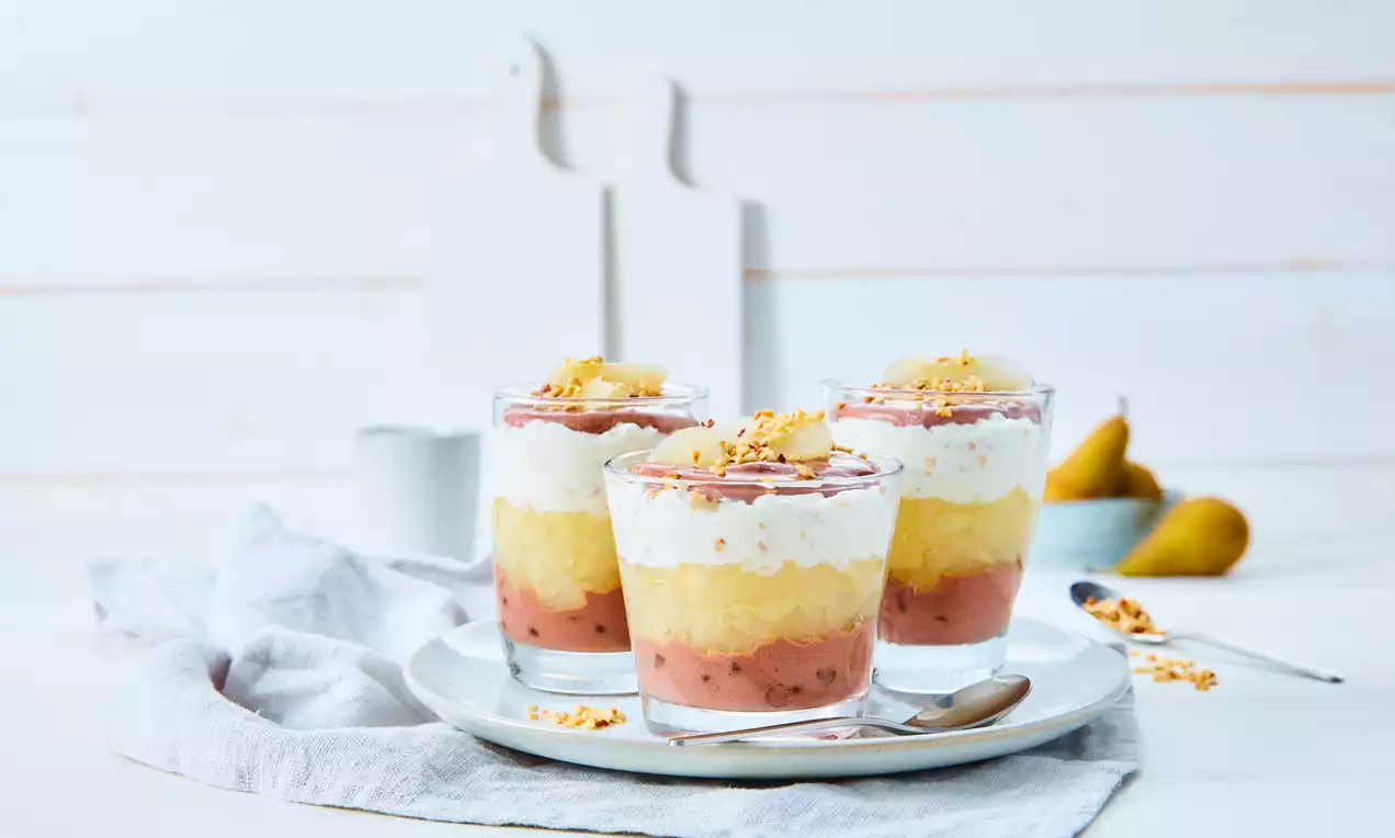 Schichtdessert aus rosa Püree, gelber Birnenmasse und weißer Creme in Gläsern, garniert mit Streuseln, auf weißem Teller mit Stoffserviette